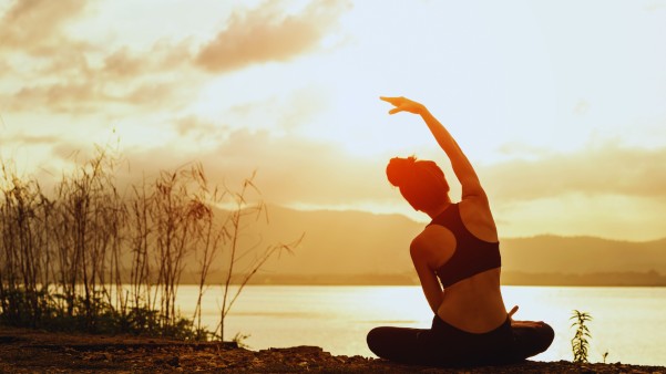 Photo of someone sitting cross legged in front of a lake at sunset or sunrise, doing stretches.