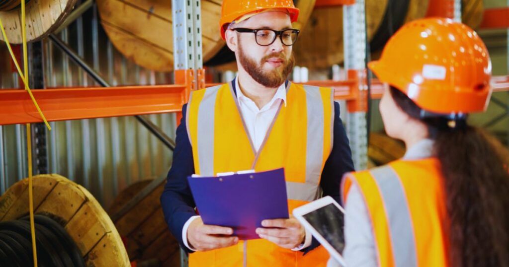 A person in an orange high visibility safety vest and hard hat is briefing another person in a warehouse setting
