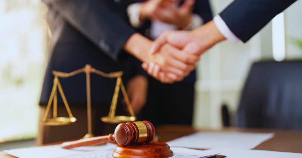 Two people in suits shake hands across a desk, on the desk is a pair of legal scales and a gavel